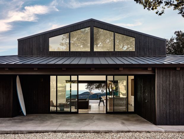 A modern single-storey residence with a dramatic raised gabled volume, dark vertical timber cladding, and a broad low-pitched metal roof. Large clerestory glazing fills the upper facade, while full-height glazed sliding doors open onto a concrete terrace or covered outdoor area. The interior is visible through the glazing, with a minimalist living space and a distant coastal or ocean view. A surfboard rests against the dark facade.
