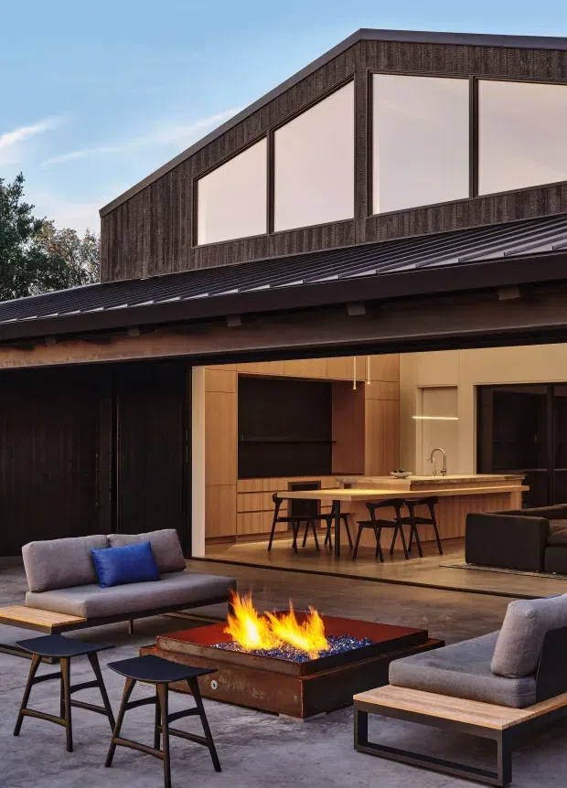 A contemporary two-storey residence with a dark textured upper facade, expansive rectangular glazing, and a low-pitched metal roof. The open ground level connects an outdoor lounge and fire-pit terrace with a warm timber-lined kitchen and dining area. Dark-framed glazing, timber cabinetry, a long island with stools, grey upholstered seating, and a linear gas fire create a refined indoor-outdoor entertaining space.