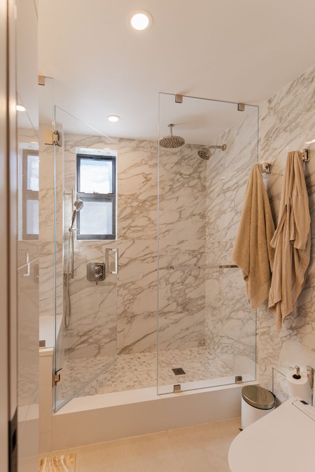 A compact contemporary bathroom centred on a glass-enclosed walk-in shower over a bathtub. The shower and surrounding walls are clad in dramatic white and beige veined marble-look stone, with brushed metal fixtures, a rainfall showerhead, handheld shower, recessed ceiling lights, mirrored surfaces, and warm beige towels. A partial white vanity and round vessel basin are visible in the foreground.