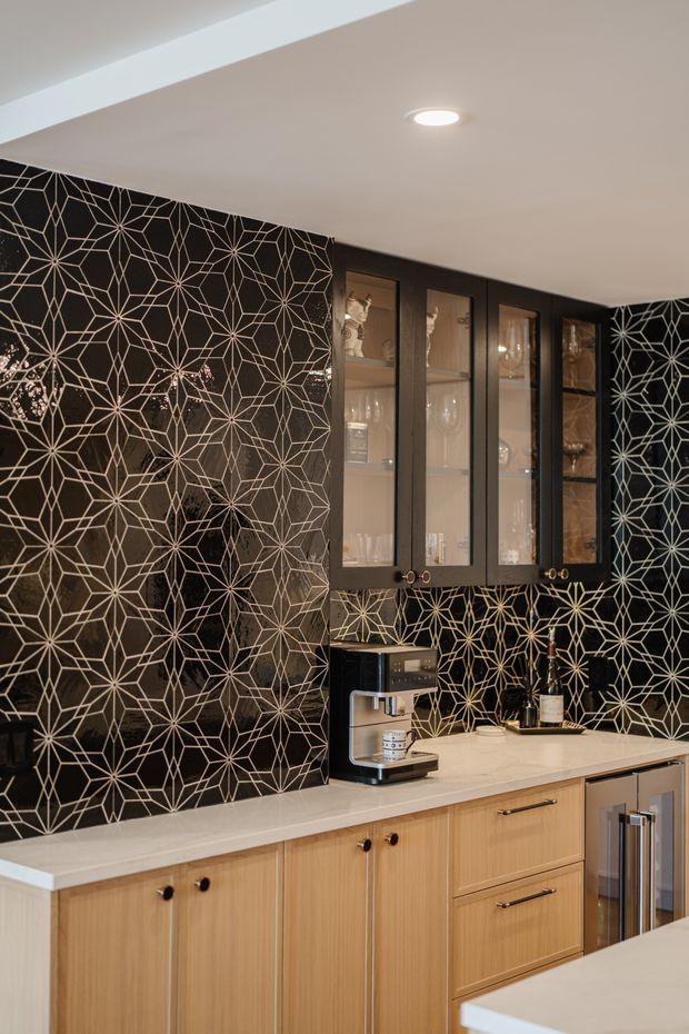 A sophisticated home bar or beverage station with light wood base cabinetry, dark framed glass-front upper cabinets displaying glassware, and a white countertop. The dramatic black geometric backsplash and side wall feature an intricate metallic starburst pattern. A compact coffee machine and wine bottle sit on the counter, with integrated beverage refrigeration at the right. Recessed ceiling lighting provides warm illumination.