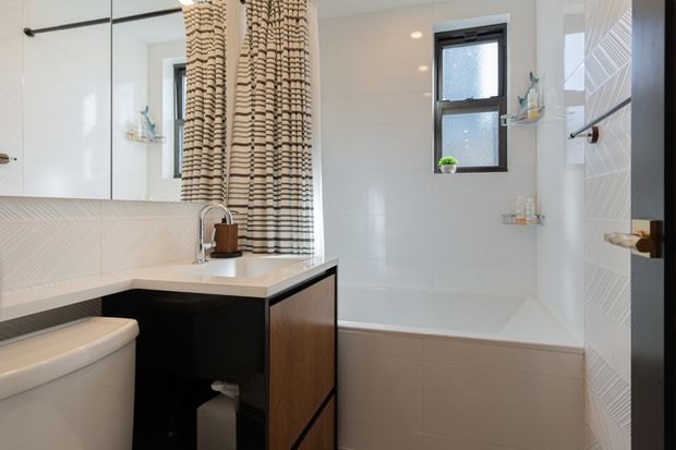 Bright contemporary bathroom with a white bathtub beneath a black-framed window, a floating dark vanity with a white benchtop and integrated basin, and a partially visible toilet. White geometric wall tiles, recessed shelving, chrome tapware, and a textured neutral shower curtain add detail. The space has a clean, compact, high-contrast fit-out with warm timber accents.