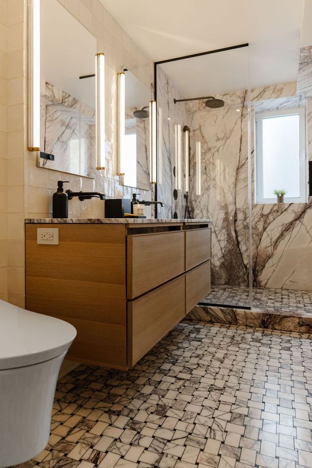 A refined contemporary bathroom featuring a floating timber vanity with a marble benchtop, wall-mounted black tapware, large marble-look wall panels, and a frameless glass walk-in shower. The floor and shower base are finished in small irregular white and grey stone tiles. Vertical illuminated bathroom mirrors and slim brass-toned details add a luxurious accent, while a freestanding white toilet is partially visible in the foreground.