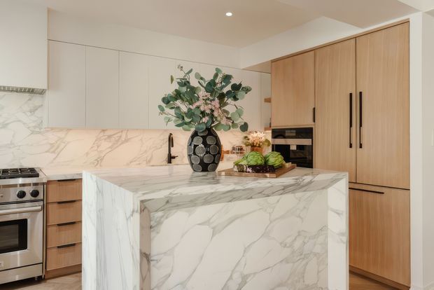 A bright contemporary kitchen centred on a large waterfall island clad in dramatic white veined stone. The room features pale timber full-height cabinetry, integrated dark appliances, white handleless upper cabinetry, a matching stone splashback, stainless steel cooking appliances, and a black sink tap. Decorative flowers and produce sit on the island, adding warmth to the otherwise refined neutral palette.