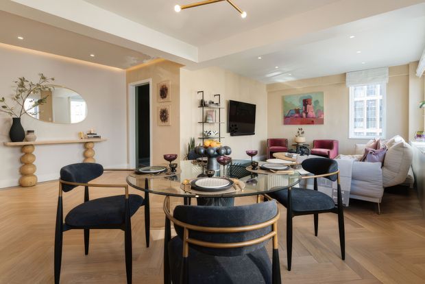 A bright, refined open-plan dining and living area. The foreground features a round glass dining table set with place settings and surrounded by dark upholstered chairs with curved timber-toned frames. Behind it, a slim console with sculptural wooden supports sits beneath a round mirror, while the adjoining living area includes a pale sectional sofa, burgundy accent chairs, a television, shelving, artwork, and large windows. Recessed lighting and a slim linear pendant provide illumination, with warm timber flooring throughout.