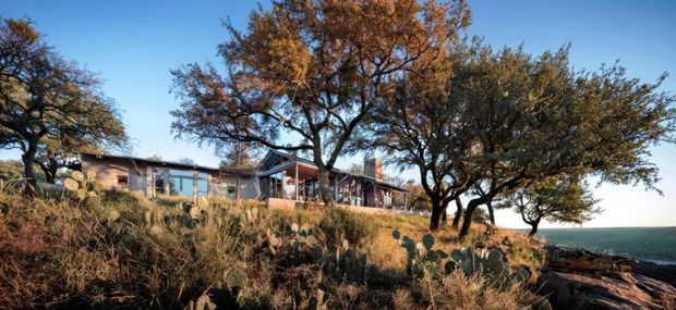 Wide exterior view of a low, elongated modern residence set among mature trees and dry native landscaping on a rocky, elevated site. The building has extensive glazing, dark timber or metal elements, and a horizontal roofline, with open water or a distant horizon visible beyond the property.
