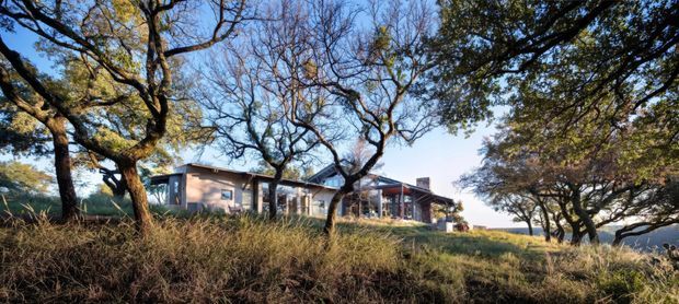 Wide exterior view of a low, contemporary residential building set among mature trees and dry grasses on a gently elevated site. The structure has a largely flat or very low-pitched roof, extensive glazing, and a muted pale facade, with an open veranda or covered outdoor connection visible along the front. The scene is photographed in warm natural light with a clear sky and distant landscape beyond.
