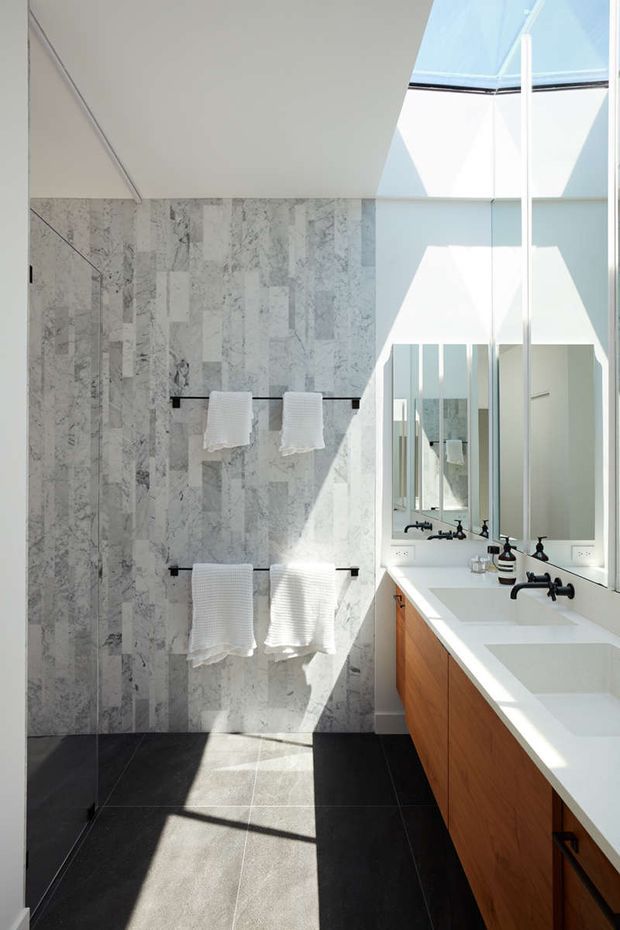 Bright contemporary bathroom with a long double vanity, white countertop, warm timber cabinetry, matte black wall-mounted tapware, and large mirrors. The walls are clad in pale grey and white veined stone tiles, while the floor uses large dark charcoal tiles. A high glazed skylight or clerestory floods the room with sunlight, creating strong geometric shadows.