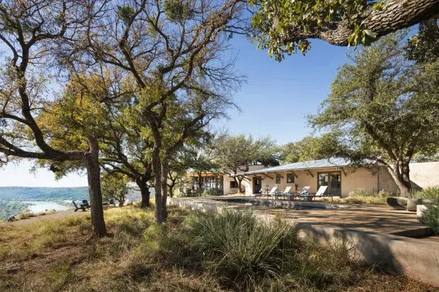 A single-storey low-profile house or lodge set among mature trees beside a broad body of water. The building has pale stucco-like walls, a shallow pitched roof with exposed-looking eaves, dark-framed windows and doors, and a long terrace with outdoor seating. Natural grasses, large trees, and the waterfront setting create a relaxed, resort-like exterior.