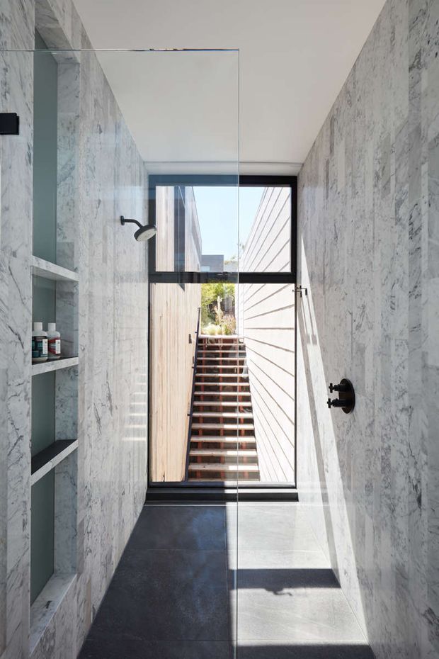 A narrow, contemporary bathroom clad in light grey-veined marble-look wall surfaces, with a frameless glass shower enclosure, black shower hardware, recessed open shelving, dark floor tiles, and a large black-framed window overlooking an exterior stairway and small planted courtyard. Strong natural light creates a clean, high-contrast atmosphere.