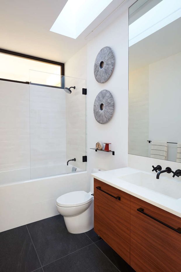 Bright contemporary bathroom with a white bathtub enclosed by a frameless glass shower screen, wall-mounted toilet, and a timber vanity with a white integrated benchtop and black wall-mounted tapware. A large mirror, skylight, recessed shelf, decorative circular wall pieces, and dark floor tiles complete the space.