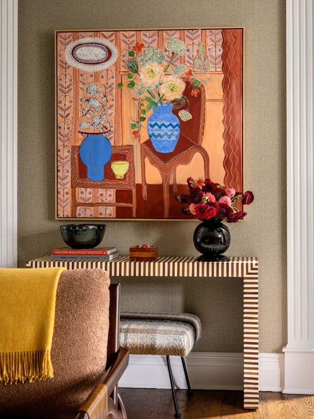 A richly styled interior vignette featuring a narrow striped console table against a textured taupe wall, topped with decorative bowls, books, a small box and a black vase of deep red flowers. A large colourful floral artwork hangs above, while a warm brown upholstered chair with mustard and blush throws partially occupies the foreground. A patterned upholstered stool sits beneath the console, with white architectural trim and dark timber flooring visible.