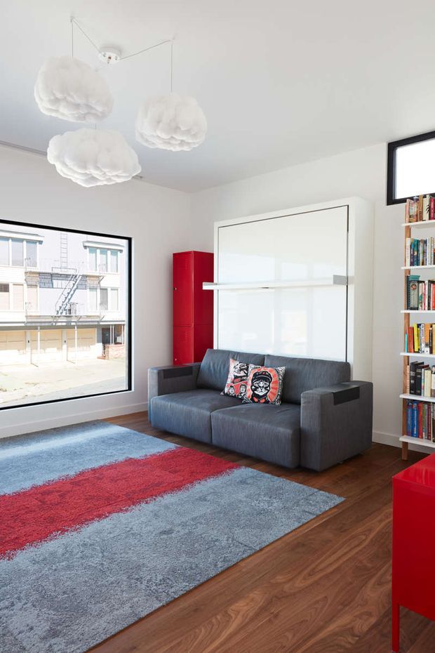 Bright, compact contemporary living room with white walls and ceiling, large black-framed windows, warm timber flooring, a dark grey sofa, and a bold blue-and-red area rug. A glossy red built-in cabinet and matching red console add strong colour, while a full-height white wall unit behind the sofa and a tall open bookshelf provide storage. Three sculptural white pendant lights hang from the ceiling.