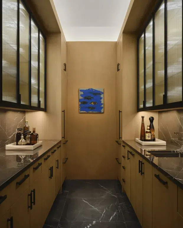 A narrow, symmetrical walk-in pantry or butler's pantry with warm timber cabinetry, dark stone benchtops and splashbacks, black-framed reeded-glass upper cabinets, black hardware, and dark marble-look flooring. Bottles sit on small trays on both counters, while a blue artwork punctuates the timber-clad rear wall. A central ceiling recess provides soft, even illumination.