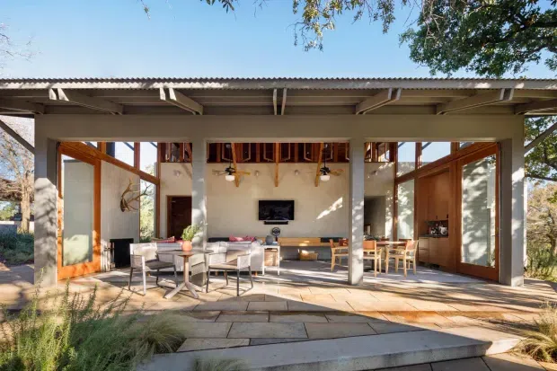 A single-storey contemporary pavilion-style home with a broad covered outdoor terrace opening directly into a living and dining area. The structure features exposed timber rafters and beams, extensive timber-framed glazing and folding or hinged doors, pale rendered walls, and a corrugated roof. Stone paving and steps form the terrace, with a sofa, dining table, television, and timber cabinetry visible inside. The setting is surrounded by mature trees and planting, creating a strong indoor-outdoor connection.
