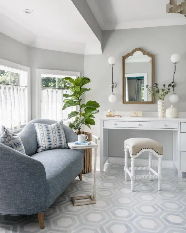 A bright, refined sitting room or dressing area with a pale grey built-in console and desk, an upholstered stool, a large blue-grey armchair, decorative mirror, twin globe wall sconces, patterned geometric flooring, and abundant natural light from two windows. A tall indoor fiddle-leaf fig adds greenery, while the palette remains soft grey, white, and muted blue.