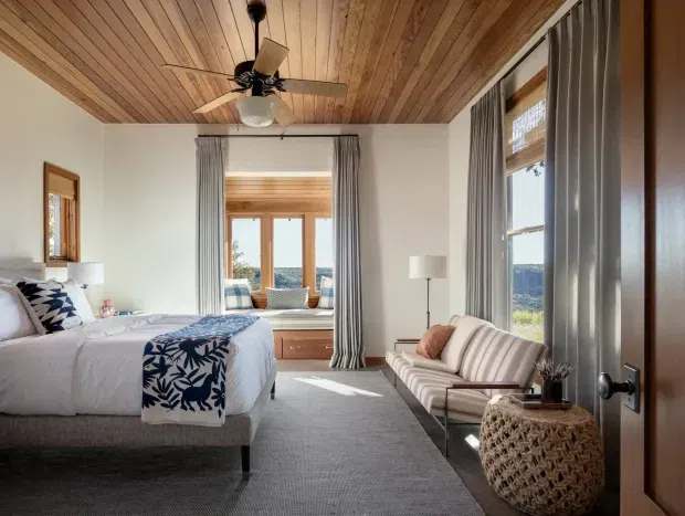 A bright, well-appointed bedroom with a large bed, striped upholstered seating, woven side table, and expansive windows. The room features a warm timber-plank ceiling with an integrated ceiling fan, white walls, grey carpeting, full-height grey curtains, and timber-framed glazing overlooking a wooded landscape. A built-in window seat and timber cabinetry add warmth, while blue-and-white patterned textiles provide a coastal accent.