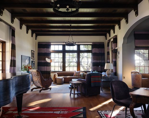 A spacious, richly furnished living room with dark exposed timber ceiling beams, cream plaster walls, large divided-light windows, and an arched opening. The room contains a grand piano, leather and upholstered seating, patterned rugs, side tables, lamps, and layered striped curtains. Warm natural light highlights the dark timber flooring and traditional architectural detailing.