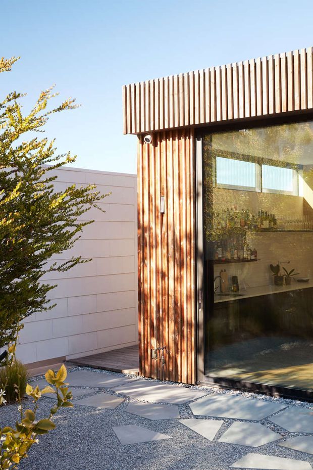 Exterior view of a contemporary building corner clad in narrow vertical timber battens, with a large dark-framed glazed opening revealing a compact interior bar or kitchenette. The facade sits beside a pale horizontal-panel wall and a landscaped gravel courtyard with irregular concrete stepping slabs. Bright, clear daylight emphasizes the warm timber and crisp geometric composition.