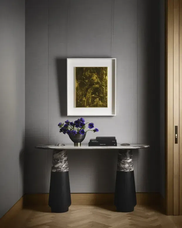 A refined interior entryway vignette with a slim marble console table supported by two sculptural dark pedestal bases. The console is styled with a black vase of purple flowers, stacked books, and a small decorative object. Behind it is a charcoal-grey vertically panelled wall with a framed abstract artwork, while a timber-lined doorway or wall feature is visible at right. The floor is warm timber parquet laid in a herringbone pattern. The composition is intimate, sophisticated, and dramatically lit.