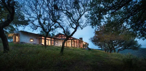 A low, modern single-storey house is nestled into a gently sloping, heavily wooded site. The darkening blue-hour scene shows broad glazed openings glowing warmly from within, a low horizontal roofline, and natural-toned walls framed by mature trees. A distant blue horizon is visible beyond the property.