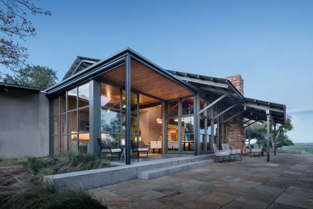 Exterior view of a contemporary single-storey pavilion-style home addition with extensive floor-to-ceiling glazing, dark structural framing, a projecting pitched roof, and a warm timber-lined soffit. The building opens onto a broad stone-paved terrace with outdoor seating, while landscaped planting and a distant water-like horizon are visible beyond. A brick chimney and the original darker rendered structure appear behind the glazed extension.
