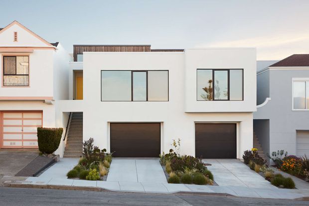 Street-facing view of a modern multi-level townhouse or attached residential development. The central white-rendered home has a clean rectilinear facade, large dark-framed windows, two integrated dark garage doors, a recessed entry stair, rooftop timber screening, and low-maintenance landscaped planting along concrete driveways. Traditional neighbouring homes are visible on either side.