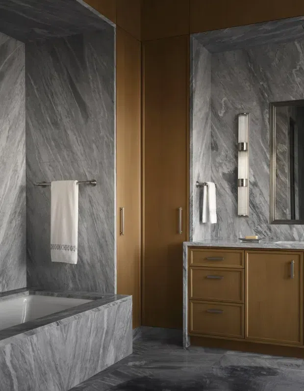 A sophisticated bathroom featuring dramatic grey veined stone slabs on the walls, floor, bathtub surround, and vanity benchtop. Warm brown wood cabinetry and tall storage doors provide contrast, while integrated towel rails, vertical wall lighting, and a framed mirror complete the refined design.