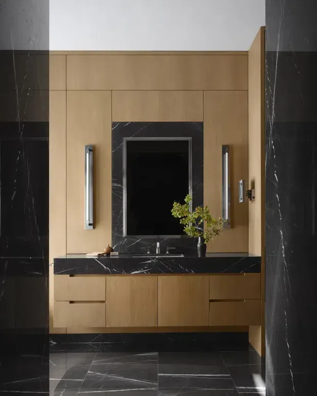 A refined bathroom vanity area with warm wood cabinetry, a black marble benchtop and backsplash surround, integrated basin, framed mirror, and vertical wall lights. Large-format dark marble-look tiles cover the surrounding walls and floor, creating a high-contrast, luxurious composition.