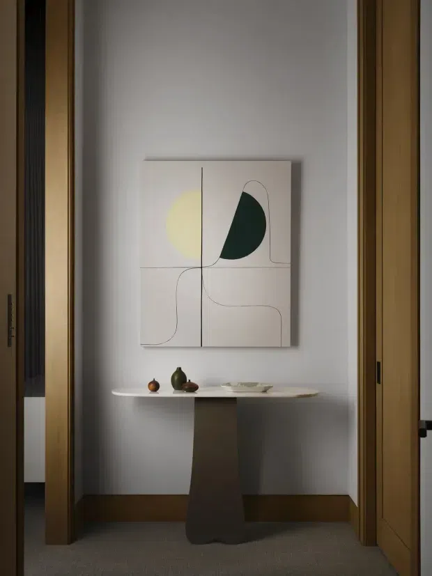 A softly lit residential hallway or entry alcove framed by warm timber door jambs, featuring a slim sculptural console table with a central pedestal, small decorative vessels, and minimalist abstract artwork above. The walls are light neutral, with dark timber skirting and a muted carpeted floor. The composition has a refined, restrained contemporary feel.