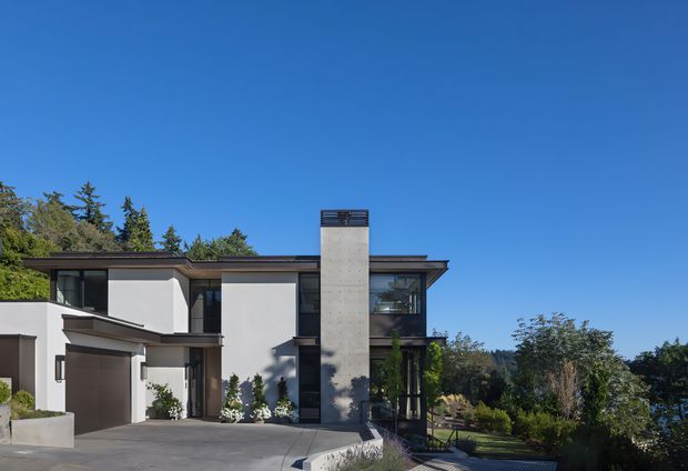 Exterior photograph of a contemporary multi-level residence with crisp white rendered walls, dark roof and window framing, broad glazing, and a prominent tall raw concrete chimney or vertical feature. The home opens onto a paved driveway and landscaped garden, with mature trees and a clear blue sky surrounding the property.