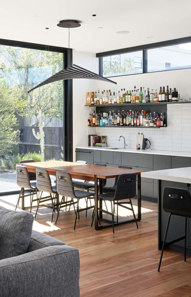 Bright open-plan kitchen and dining area with a built-in home bar, dark cabinetry, white tiled splashback, timber dining table, upholstered chairs, and expansive black-framed glazing opening to a leafy outdoor setting. A sculptural linear pendant light hangs above the dining table, while a grey sofa is partially visible in the foreground.