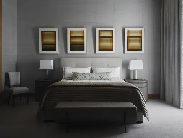 A refined, symmetrical bedroom with a dark grey textured feature wall, upholstered bed, layered white bedding, and a long charcoal quilt. Four framed artworks are arranged above the bed, flanked by bedside tables and matching lamps. A dark upholstered bench sits at the foot of the bed, with full-height grey curtains and soft carpet completing the calm, moody interior.