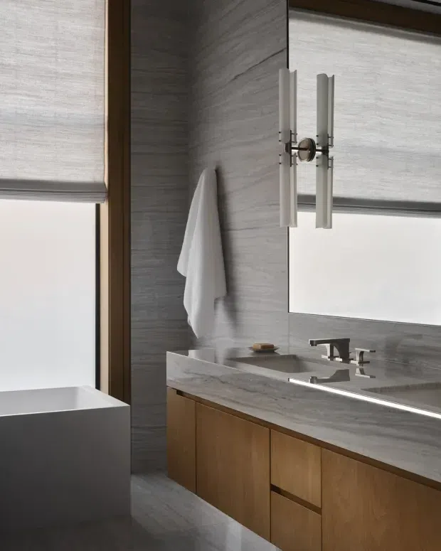A refined contemporary bathroom with a long timber vanity featuring integrated cabinetry and a pale grey veined stone benchtop and backsplash. The vanity includes a double-sink arrangement with sculptural metal tapware, a large wall mirror, and vertical contemporary wall sconces. A freestanding rectangular bathtub is visible beside a tall window with translucent roller blinds. Grey stone-look wall and floor finishes, warm timber joinery, and soft natural light create a calm, luxurious atmosphere.
