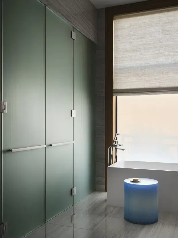 A contemporary bathroom featuring tall frosted glass shower or storage doors with slim metal hinges and handles, grey textured wall cladding, and grey stone-look floor tiles. A freestanding white bathtub sits beside a large window with a woven roller blind. A small translucent blue cylindrical stool or side table is positioned in front of the tub. The room has a refined, cool-toned, minimalist appearance with soft natural light.