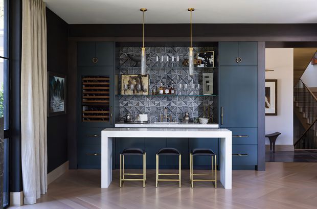 A sophisticated built-in home bar is integrated into a dark blue-grey joinery wall. The bar features a patterned tiled backsplash, open display shelving, wine storage, a pale stone-look counter, brass-accented pendant lights, and three bar stools. Timber flooring, dark walls, full-height curtains, and a glimpse of a stair hall create a refined, moody interior.