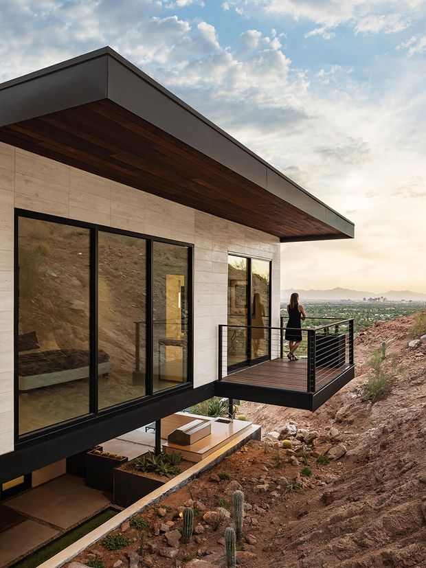 Exterior architectural photograph of a contemporary angular house on a steep rocky hillside. The building features a projecting flat roof with a dark timber soffit, pale rectangular wall cladding, extensive black-framed floor-to-ceiling glazing, and a projecting balcony with black horizontal cable or metal railings. A lower terrace with built-in planters is visible beneath the cantilevered volume, with a broad valley and distant mountains beyond.