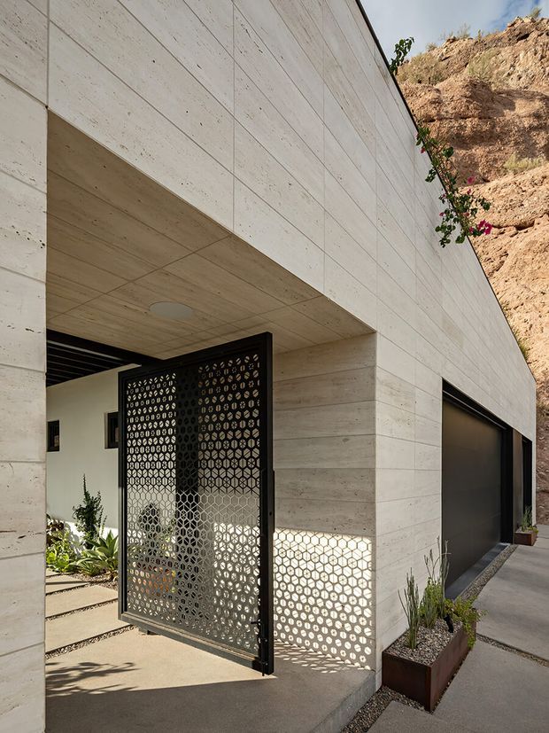 Modern hillside residential exterior with a monolithic pale concrete facade, recessed entry, perforated black metal pivot gate, and an integrated dark garage door. The building is cut into a rocky, planted slope, with patterned sunlight cast through the entry screen onto the concrete wall and paving.