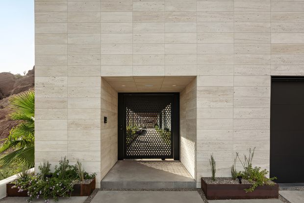 Modern exterior entry facade clad in large light beige stone or concrete panels. A deeply recessed doorway features a black perforated metal screen and dark frame, with a broad dark garage opening to the right. Linear planters with drought-tolerant landscaping flank the entrance, and patterned sunlight falls across the threshold.