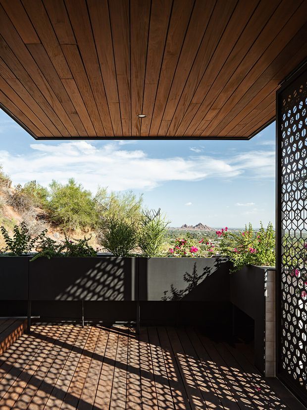 A shaded outdoor terrace or balcony with a dark timber-plank ceiling and matching timber decking. A perforated geometric metal privacy screen at the right casts patterned shadows across the floor and built-in dark cabinetry or planter edges. The open view looks onto arid landscaped terrain and distant rocky hills, with flowering plants along the terrace perimeter.