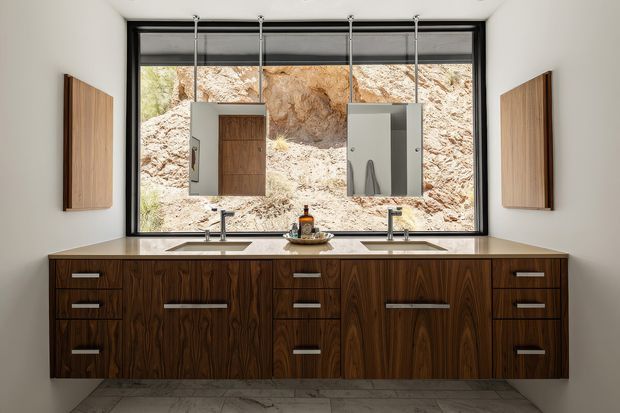 A contemporary double-vanity bathroom with a long dark timber floating cabinet, two integrated sinks, wall-mounted brushed metal taps, and matching mirrored medicine cabinets. A large black-framed window provides a rocky natural outlook, while vertical timber wall panels flank the vanity. The room has light walls and a pale stone-look floor, with a clean, refined, warm modern aesthetic.