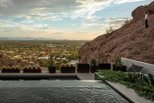 A contemporary hillside outdoor terrace overlooks a sprawling suburban valley. The foreground features a long rectangular reflecting pool or swimming pool, pale concrete paving, dark raised planters with sculptural desert plants, and a low white retaining wall. A steep exposed rocky hillside rises immediately behind the terrace, with a person standing on the slope. The scene has expansive mountain and city views beneath a partly cloudy sky, with a muted natural palette and refined landscape design.
