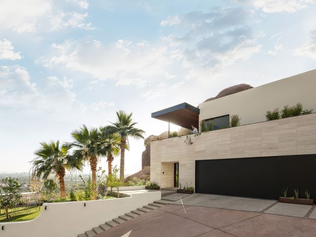 Exterior view of a contemporary multi-level residence built into a rocky, landscaped hillside. The home features pale horizontal stone or masonry cladding, a smooth cream upper volume, dark recessed garage doors, broad concrete paving, a projecting flat-roof canopy, and integrated outdoor steps and terraces. Palm trees and a distant valley are visible around the property.