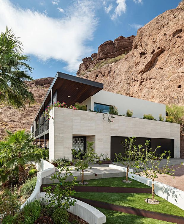 Exterior view of a contemporary multi-level residence built into a rugged desert canyon. The home has pale stone-clad walls, dark-framed glazing, a broad projecting roof with a timber-lined soffit, upper terraces with planted edges, and a dark garage door. Curved white retaining walls, manicured lawn, palms, and desert planting form the landscaped foreground, with dramatic exposed rock cliffs directly behind the house.