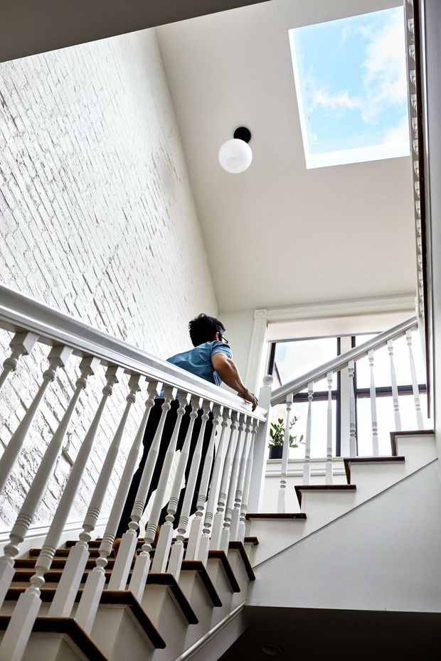 Bright interior view looking up a stairway and landing in a multi-level home. The staircase has white painted turned balusters and handrails with dark timber stair treads, opening toward a landing with large windows and a roof skylight. A textured white-painted brick wall, simple globe pendant, and a person ascending the stairs are visible.