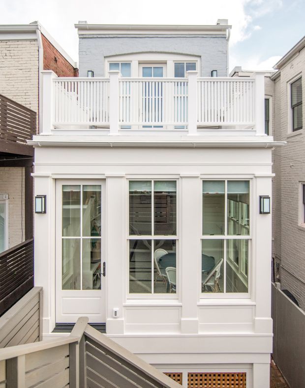 Front-facing view of a narrow multi-storey urban townhouse facade. The predominantly white-painted exterior features a projecting upper balcony with vertical balustrades, tall glazed doors and windows, decorative horizontal trim and pilaster-like framing, wall-mounted exterior lights, and a lower-level screened opening. Adjacent tightly spaced brick buildings and privacy fencing emphasize the compact urban setting.