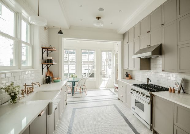 Bright, elongated traditional-style kitchen with pale grey built-in cabinetry, white subway-tile walls, white benchtops, and a large freestanding stainless-steel range cooker. The room features tall multi-pane windows, glazed doors at the far end, a farmhouse-style sink, open shelving, pendant lighting, and a patterned tiled floor. A small breakfast nook is visible beyond the main kitchen zone.