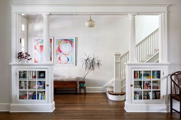 Bright, refined transitional interior showing a formal hallway or landing with white classical columns, built-in bookcases, decorative trim, hardwood flooring, artwork, and a staircase rising at the right. A small sitting area and potted plant are visible beyond the opening.