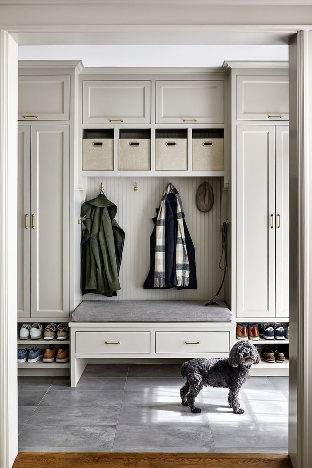 A built-in mudroom entryway with symmetrical light-grey painted cabinetry, raised-panel doors, brass pulls, open cubbies, woven storage baskets, a central upholstered bench, coat hooks, and lower shoe shelving. Large grey floor tiles contrast with a timber threshold, while coats, scarves, hats, shoes, and a dog provide everyday context. The joinery has a polished traditional feel with clean, balanced detailing.