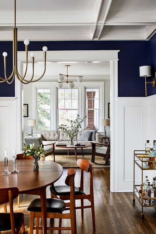 Refined open-plan dining and living interior with deep navy walls, white decorative wall panelling and ornate coffered ceilings. A mid-century wooden dining table and chairs occupy the foreground, while a seating area with a sofa, armchairs and coffee table sits beyond a wide cased opening. Brass and glass lighting, a drinks trolley, timber flooring and layered traditional trim create a sophisticated, warmly styled setting.