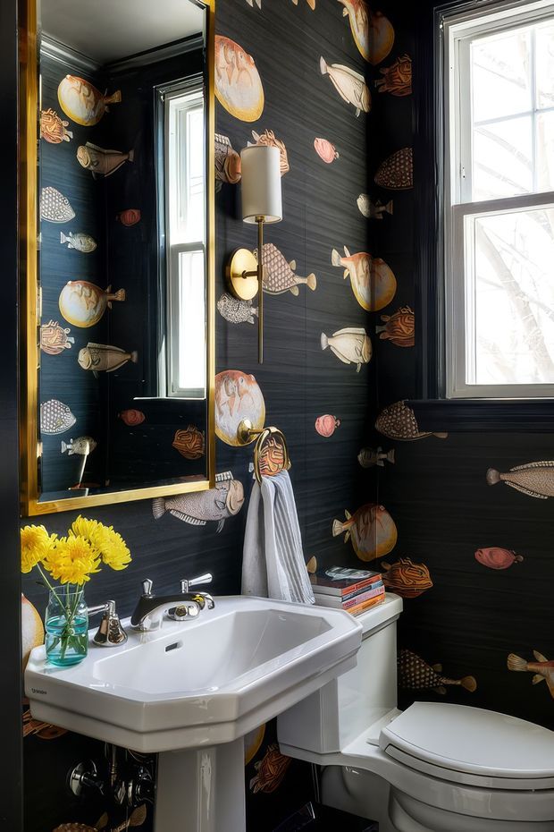 A compact, dramatically styled bathroom with dark fish-pattern wallpaper covering the walls, black trim, and a tall white-framed window. A large gold-framed mirrored cabinet, white pedestal sink with chrome tapware, toilet, wall sconce, and small floral arrangement create a playful vintage-inspired interior.